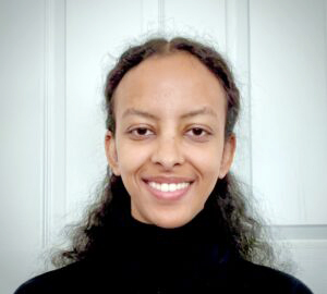 A person with long, curly hair and a black turtleneck smiles at the camera, standing in front of a white paneled door at the Lutheran Church Aldie Virginia.