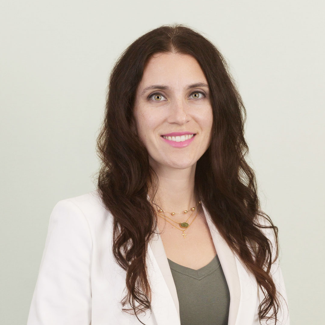 Woman with long brown hair wearing a white blazer over a green top, smiling at the camera. She has light skin, layered gold necklaces, and stands against a plain, light-colored background at Lutheran Church Aldie Virginia.