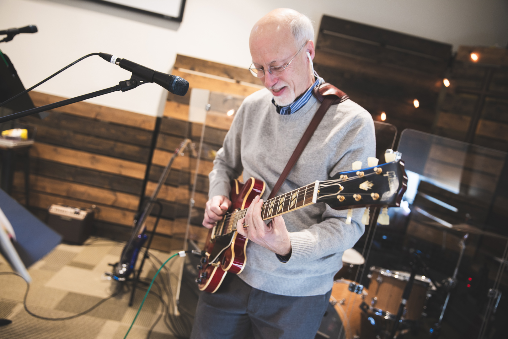 An older man with glasses and earbuds plays an electric guitar and smiles while standing near a microphone in a music room with wooden walls and drums in the background.