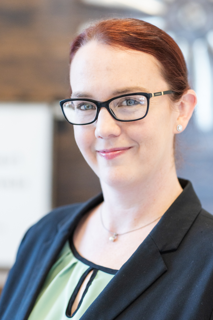 A woman with red hair in a bun, wearing glasses, a black blazer, and a light green blouse, smiles slightly while standing indoors with a blurred background.