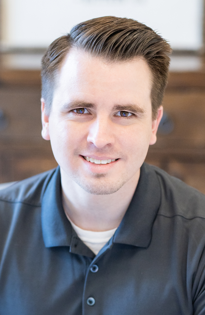 A young man with short brown hair and light skin, wearing a dark gray polo shirt, smiles at the camera. The background is softly blurred, featuring wooden furniture.