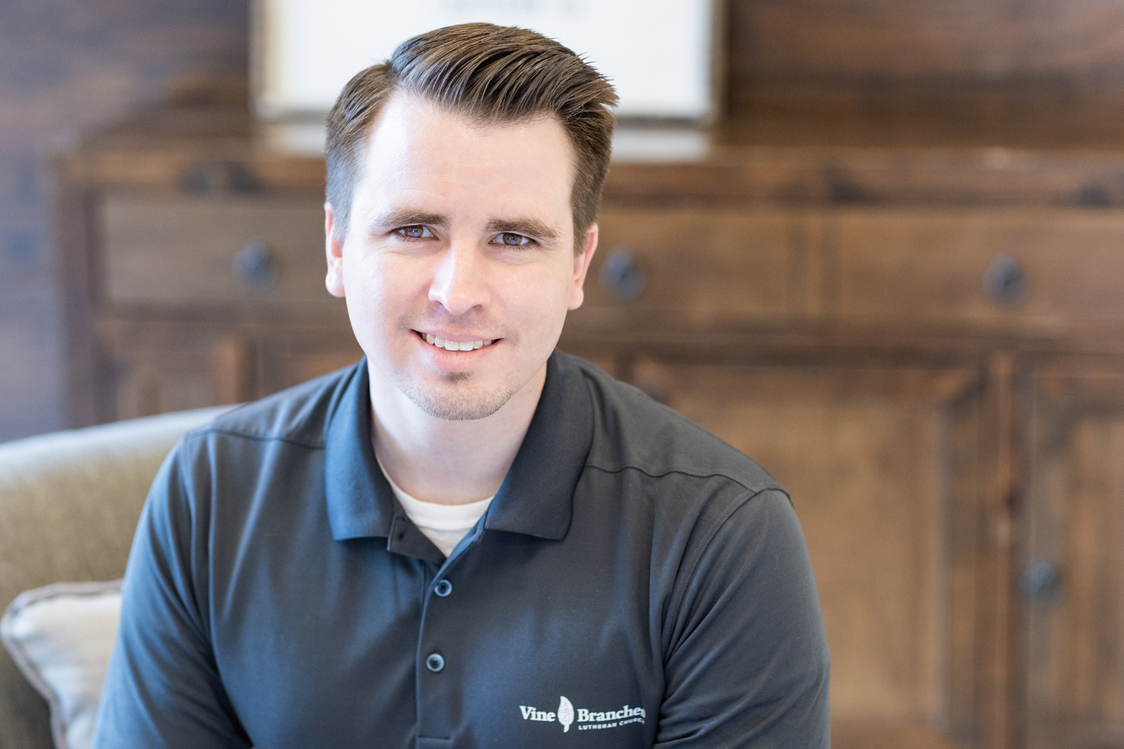 A young man with short brown hair, wearing a dark gray polo shirt that reads Vine Branch sits and smiles at the camera. The background features a wooden cabinet.