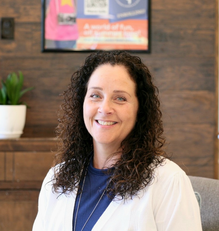 A woman with curly dark hair, wearing a white cardigan and navy shirt, smiles while sitting indoors. There is a plant and a blurred poster in the background.